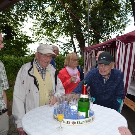 Gruppe älterer Menschen bespricht sich um einen Tisch mit Sektflasche und Gläsern im Freien unter Bäumen.