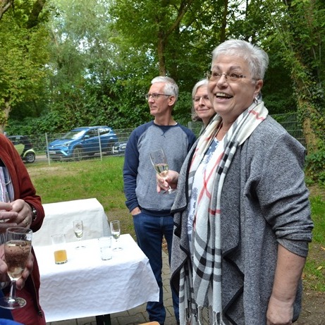 Gruppe von Personen bei einem Treffen im Freien, die Getränke halten und sich unterhalten, umgeben von Bäumen.