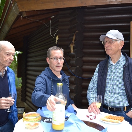 Drei Männer stehen an einem Tisch im Freien und unterhalten sich, während Getränke und Snacks auf dem Tisch stehen.