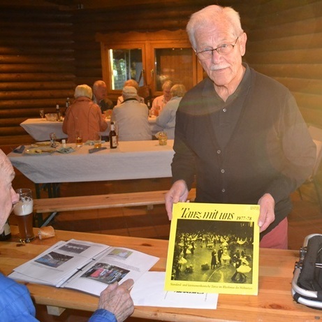 Älterer Mann hält eine Zeitschrift mit dem Titel „Tanz mit uns“ in einem hochgelegenen Raum mit mehreren Gästen.