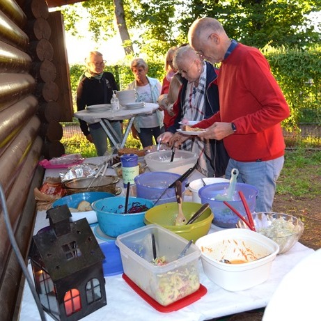 Personen stehen an einem Buffet mit verschiedenen Schalen und Töpfen auf einem Tisch im Freien, umgeben von Natur.