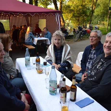 Sechs Personen sitzen an einem Tisch unter einem Zelt, Getränke stehen bereit, im Hintergrund sind weitere Gäste und Fahrräder.