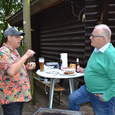 Zwei Männer unterhalten sich am Tisch im Freien, umgeben von Bäumen, mit Getränken und Grillgut auf dem Tisch.