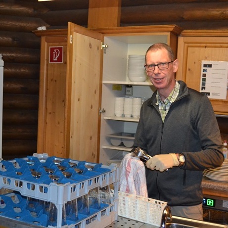Mann mit Handschuhen reinigt Gläser in einer holzverkleideten Küche mit Geschirr und Utensilien.