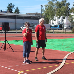Zwei Personen in roten T-Shirts sprechen auf einem Sportplatz mit grüner Matte und Tribüne im Hintergrund.