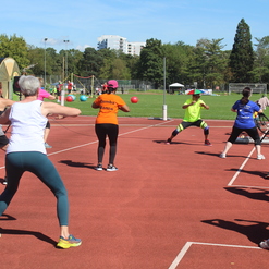 Gruppe von Frauen in Sportbekleidung bei einer Fitnessübung auf einem roten Sportplatz, mit Bällen und Musikanlage im Hintergrund.