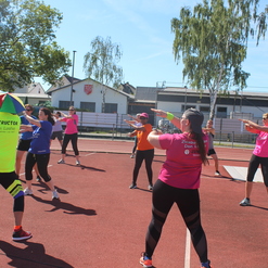 Zehn Teilnehmerinnen in Sportkleidung machen Zumba-Übungen auf einem roten Sportplatz unter blauem Himmel.