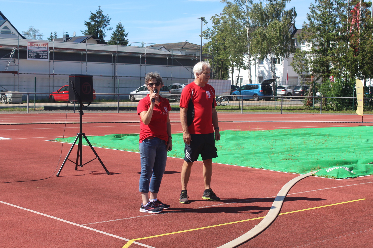 Zwei Personen in roten T-Shirts sprechen auf einem Sportplatz mit grüner Matte und Tribüne im Hintergrund.