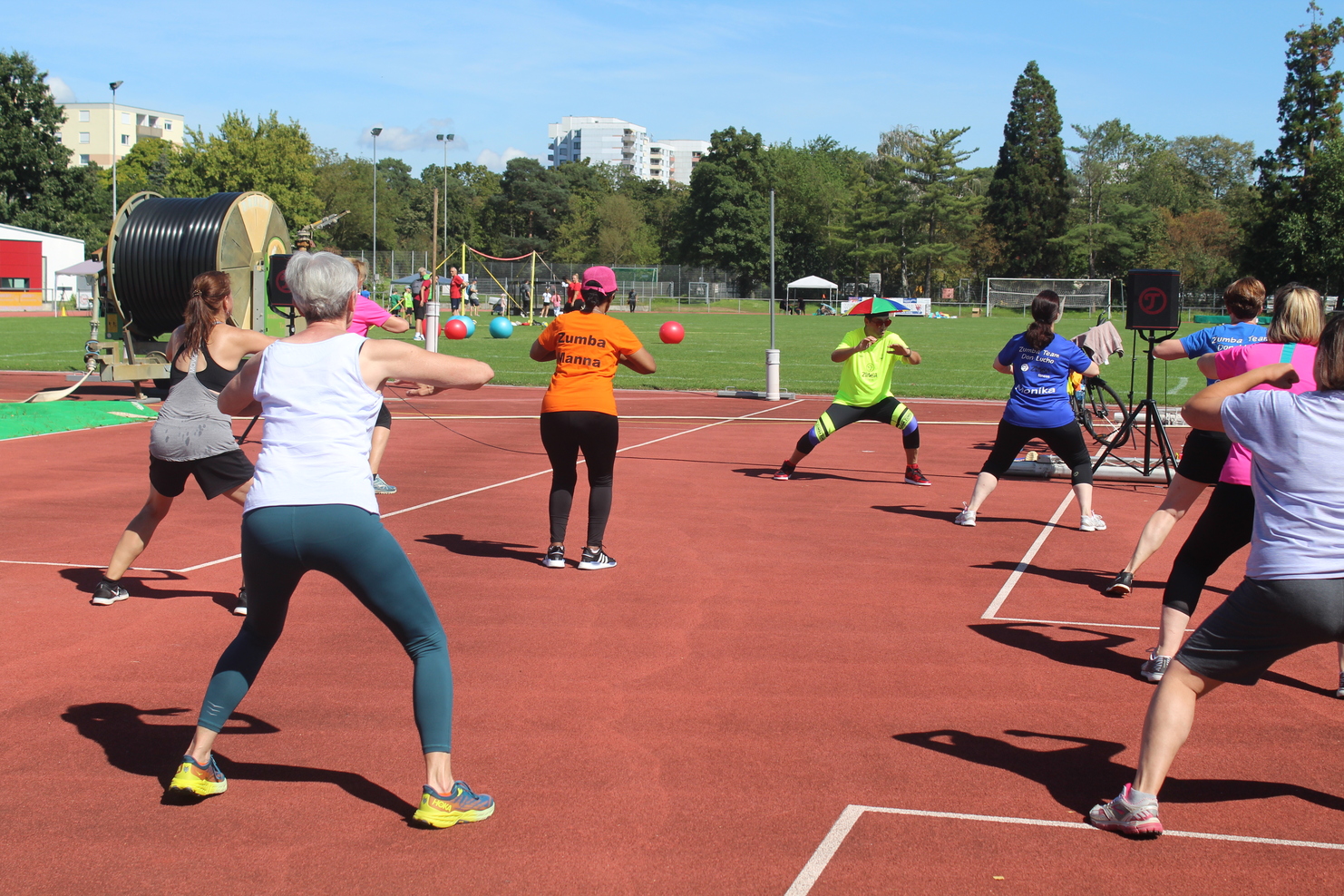 Gruppe von Frauen in Sportbekleidung bei einer Fitnessübung auf einem roten Sportplatz, mit Bällen und Musikanlage im Hintergrund.