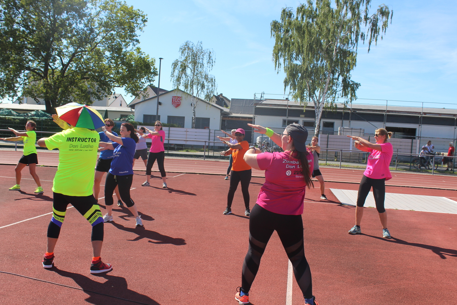 Zehn Teilnehmerinnen in Sportkleidung machen Zumba-Übungen auf einem roten Sportplatz unter blauem Himmel.
