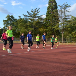Gruppe von Frauen in Sportkleidung trainiert auf einem roten Sportplatz unter blauem Himmel.
