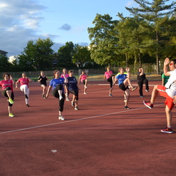 Gruppe von Frauen und einem Mann beim Training auf einem Sportplatz, alle heben die Knie in die Luft.