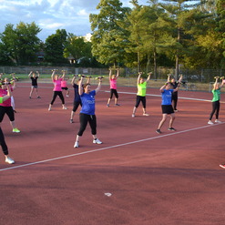 Gruppe von Frauen und einem Mann beim Fitness-Training mit Gewichten auf einem roten Sportplatz unter blauem Himmel.