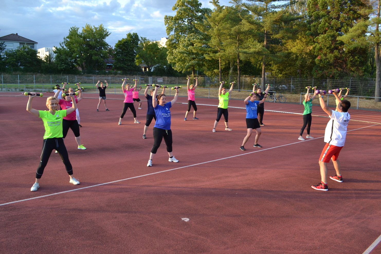 Gruppe von Frauen und einem Mann beim Fitness-Training mit Gewichten auf einem roten Sportplatz unter blauem Himmel.