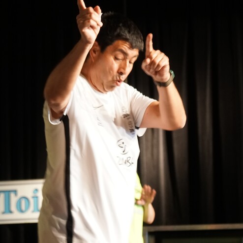 Mann im weißen Shirt und roten Shorts tanzt mit erhobenen Händen auf einer Bühne mit schwarzem Hintergrund.