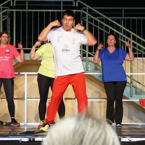 Gruppen von Zumba-Tänzern in bunten T-Shirts führen einen aktiven Tanz auf einer Bühne vor Publikum auf.