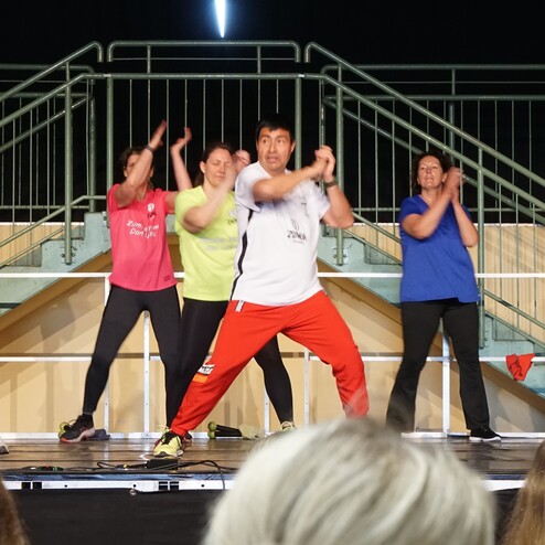 Gruppentanz von Zumba-Teilnehmern auf einer Bühne, in bunten T-Shirts, energetischer Bewegungsstil.