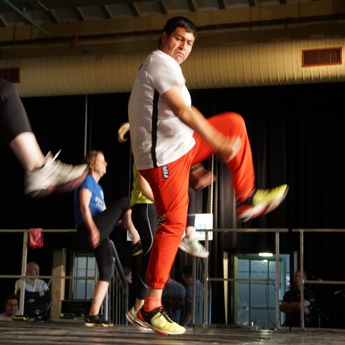 Aktive Teilnehmer bei einem Tanztraining, einer Mann hebt sein Bein, während andere im Hintergrund tanzen.