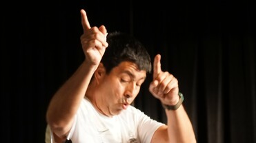 Mann in Zumba-T-Shirt tanzt energisch auf einer Bühne und hebt die Hände in die Luft, im Hintergrund eine zweite Person.