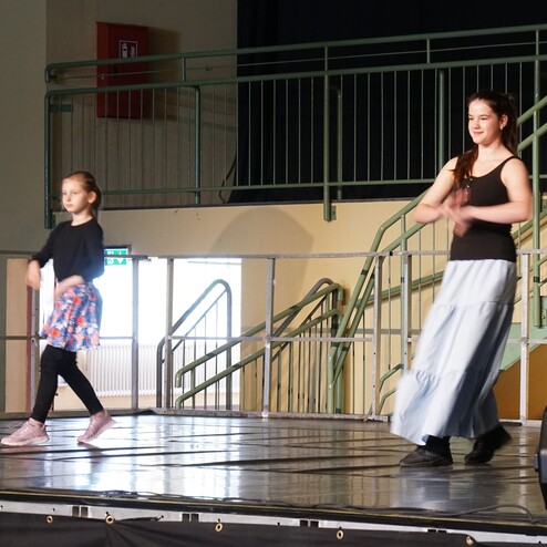 Zwei T&auml;nzerinnen f&uuml;hren eine Choreografie auf einer B&uuml;hne mit grauem Boden und einer Treppe im Hintergrund vor.