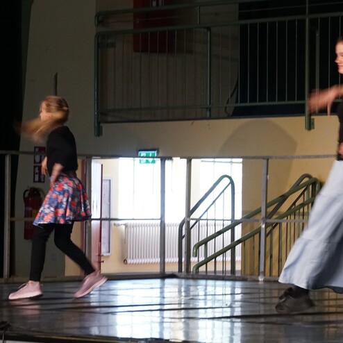 Zwei T&auml;nzerinnen auf einer B&uuml;hne in Bewegung, eine tr&auml;gt einen Rock, die andere eine lange Hose.