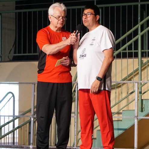 Zwei M&auml;nner stehen auf einer B&uuml;hne, einer h&auml;lt ein Mikrofon, der andere spricht, beide tragen Sportkleidung.