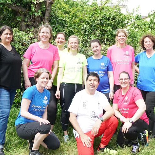 Gruppe von zehn Zumba-Mitgliedern in bunten Shirts, stehend und kniend vor einer gr&uuml;nen Hecke.
