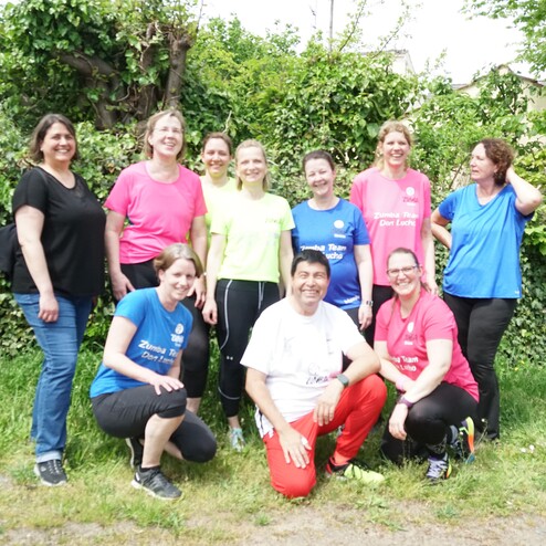 Gruppe von zehn Personen in bunten Zumba-Shirts posiert l&auml;chelnd vor einer gr&uuml;nen Hecke im Freien.