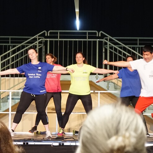 Zumba-Gruppe in Sportkleidung f&uuml;hrt synchron Tanzbewegungen auf einer B&uuml;hne aus. Zuschauer sind im Vordergrund sichtbar.