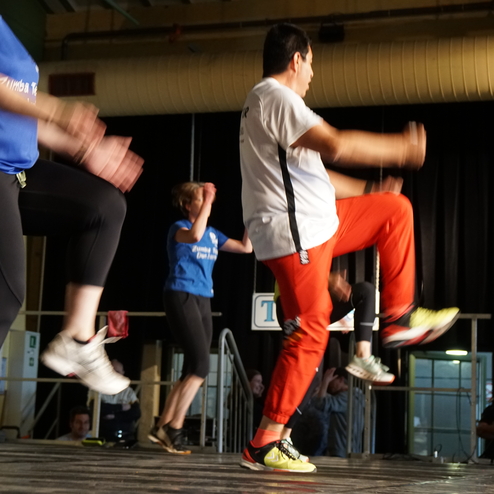 Zwei Gruppen von Personen in Sportkleidung tanzen synchron auf einer B&uuml;hne w&auml;hrend einer Zumba-Veranstaltung.