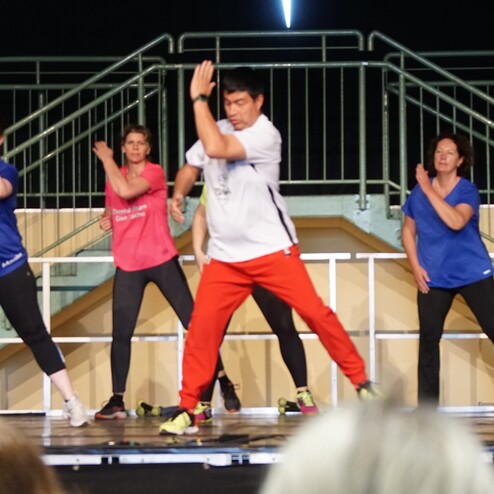 Gruppe von acht T&auml;nzern in Sportkleidung, die synchron auf einer B&uuml;hne Zumba tanzen.