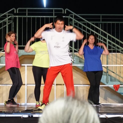 Gruppenvorf&uuml;hrung mit acht Personen in Sportkleidung, die Zumba-&Uuml;bungen auf einer B&uuml;hne ausf&uuml;hren.