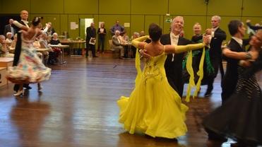 Tanzpaare in eleganter Kleidung tanzen auf einer Wettkampf-Bühne vor einem Publikum in einem festlichen Saal.
