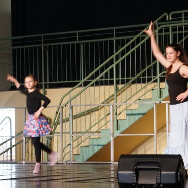 Zwei Tänzerinnen auf einer Bühne: ein Mädchen in Rock und ein Mädchen in langer Bluse, beide in aktiver Pose.