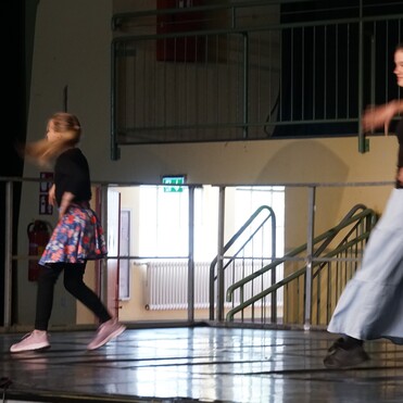 Zwei Tänzerinnen auf einer Bühne in Bewegung, eine trägt einen Rock, die andere eine lange Hose.