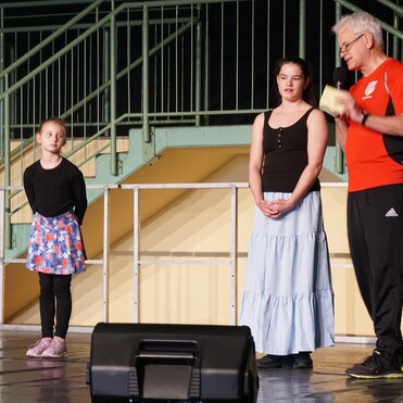 Zwei Mädchen stehen auf einer Bühne, während ein Mann in Sportkleidung mit Mikrofon spricht. Treppe im Hintergrund.