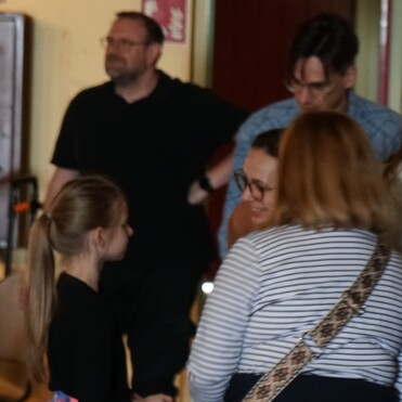 Gruppe von Menschen in einem Raum, einige im Gespräch, ein Mädchen steht im Vordergrund und hört aufmerksam zu.