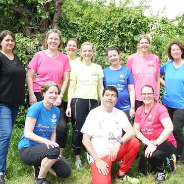 Gruppe von zehn Zumba-Mitgliedern in bunten Shirts, stehend und kniend vor einer gr&uuml;nen Hecke.