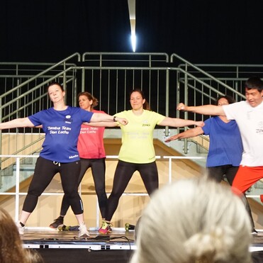 Zumba-Gruppe in Sportkleidung führt synchron Tanzbewegungen auf einer Bühne aus. Zuschauer sind im Vordergrund sichtbar.