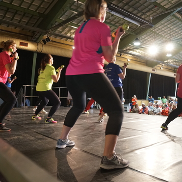Fünf Frauen in Sportkleidung führen auf einer Bühne Krafttraining mit Hanteln vor Publikum aus.