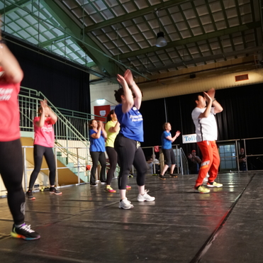 Verschiedene tanzende Personen in bunten Shirts und Sportkleidung auf einer Bühne in einem Fitness-Event.