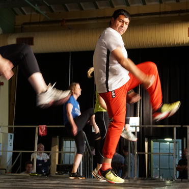 Gruppentraining mit mehreren Teilnehmern, die aktiv ihre Beine heben, auf einer B&uuml;hne in einem Sportbereich.