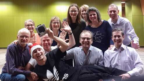 Gruppenselfie mit zw&ouml;lf Personen, die in einem Raum lachen und posieren, einige halten ein Geschenk in den H&auml;nden.