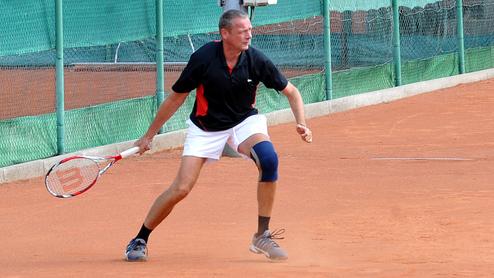 Mann mit Tennisschläger in Bewegung auf einem Sandplatz, umgeben von grünen Netz- und Platzabsperrungen.