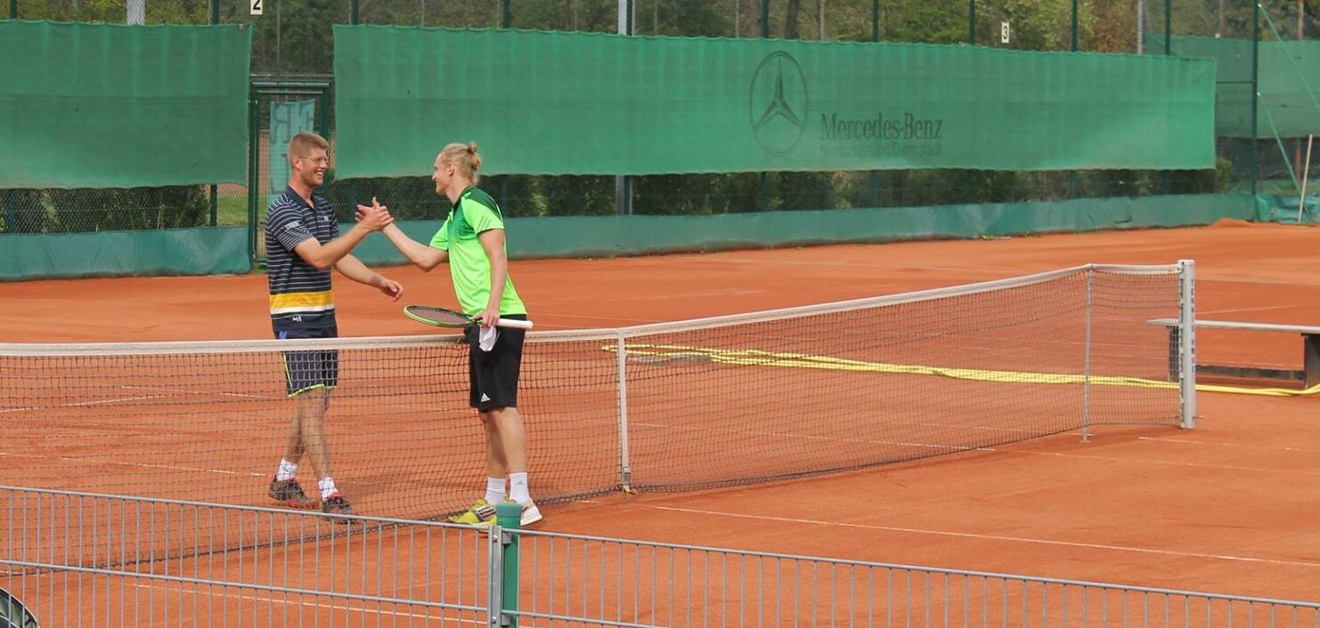 Zwei Tennis-Spieler schütteln sich am Netz die Hände auf einem Sandplatz, umgeben von grünen Zaunplanen.