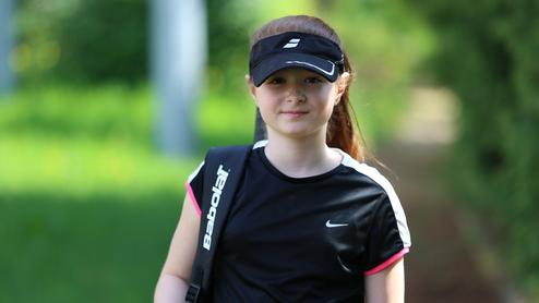 Junge im sportlichen Outfit mit Tennisrackett und Tasche, steht auf einem Weg, umgeben von grünem Gras und Bäumen.