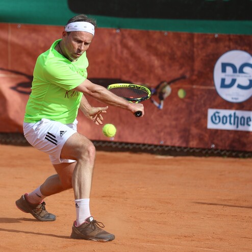 Tennisspieler in gr&uuml;nem Shirt schl&auml;gt einen Ball auf einem Sandplatz, im Hintergrund Werbebanner.