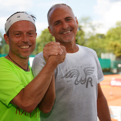 Zwei M&auml;nner, einer in gr&uuml;nem Shirt, der andere in grauem Shirt, sch&uuml;tteln sich l&auml;chelnd die H&auml;nde auf einem Tennisplatz.