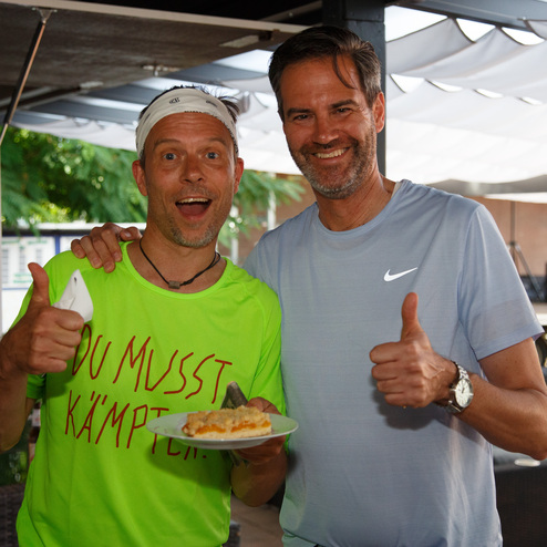 Zwei M&auml;nner stehen l&auml;chelnd nebeneinander, einer h&auml;lt einen Teller mit Kuchen und beide zeigen Daumen hoch.