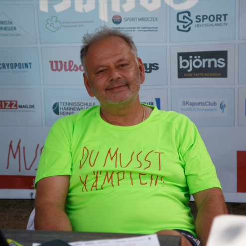 Mann mit grauen Haaren und T-Shirt in Neonfarbe mit der Aufschrift &bdquo;DU MUSST K&Auml;MPFEN!&ldquo; sitzt am Tisch.